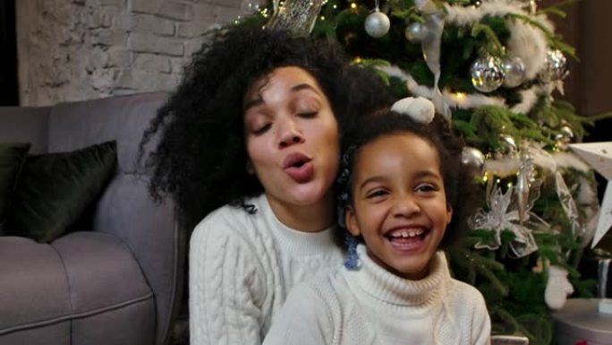 快乐微笑的非洲裔美国妈妈和小女儿祝圣诞快乐，新年快乐的肖像。家庭在家里装饰圣诞树的背景下。特写。慢动