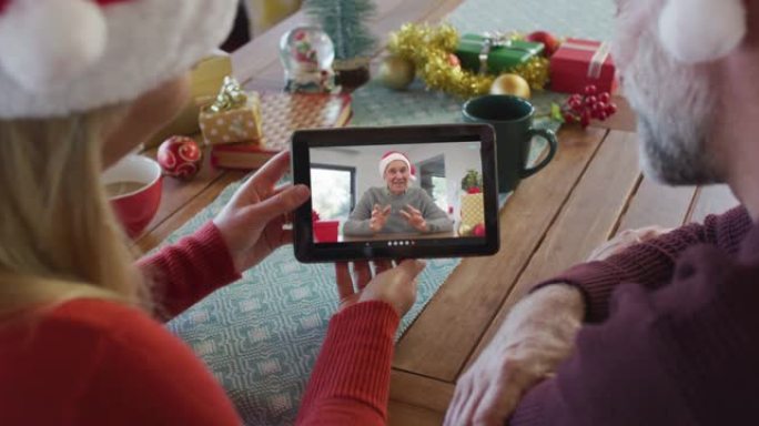 戴着圣诞老人帽子的高加索夫妇使用平板电脑进行圣诞节视频通话，屏幕上有微笑的家人