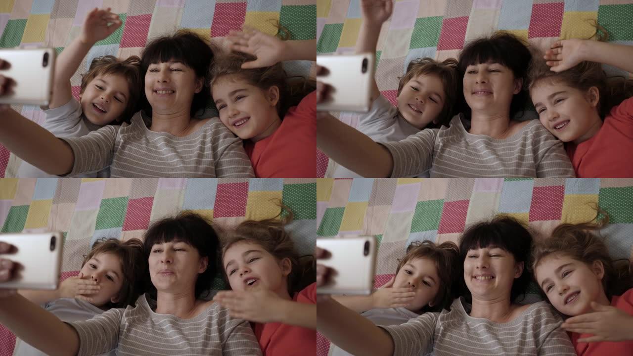 视频通话家庭自拍照。肖像母亲和孩子们在镜头前接吻。快乐的母亲和她的孩子正在床上给父亲或亲戚自拍或视频