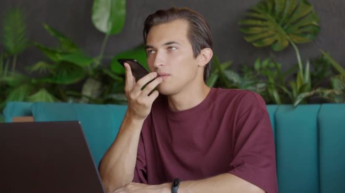 戴眼镜的英俊年轻人坐在咖啡馆的桌子旁，通过智能手机进行语音聊天。这家伙给社交网络写语音邮件。