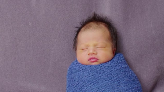 可爱的睡眠新生儿在灰色毯子背景上的蓝色弹力包裹材料中放松。亚洲新生儿婴儿睡眠舒适温暖。新生儿摄影概念