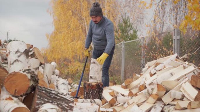 一个穿着蓝色毛衣和牛仔裤的男人在秋天的房子后院用斧头砍柴。伐木工人砍柴后会擦汗
