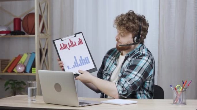 穿着方格衬衫的年轻人使用笔记本电脑和耳机在视频电话会议上聊天，显示销售图表。红发男性，留着胡须坐在家