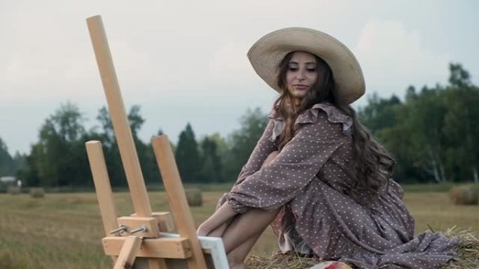 夏天日落时分，穿着太阳裙和帽子的快乐创意女艺术家在田野里坐在干草堆上，欣赏画架上的绘画，微笑着。有创