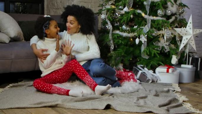 非洲裔美国妈妈和她的小女儿的肖像，表情震惊而惊讶。女人和女孩坐在沙发和装饰圣诞树附近。慢动作