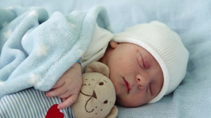初生婴儿脸部肖像的特写镜头，早年与蓝色背景上的柔软泰迪熊一起睡觉。孩子在帽子上生命的开始。婴儿，分娩