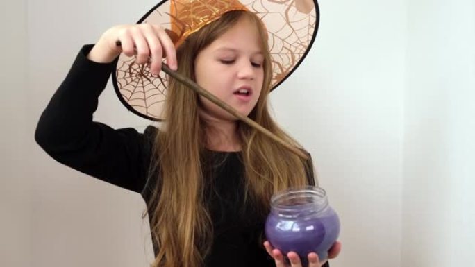 戴着橙色女巫帽子的小女孩正在制作魔法药水。孩子穿着万圣节派对的服装。孩子在杯子里搅拌着柔软的紫色物质
