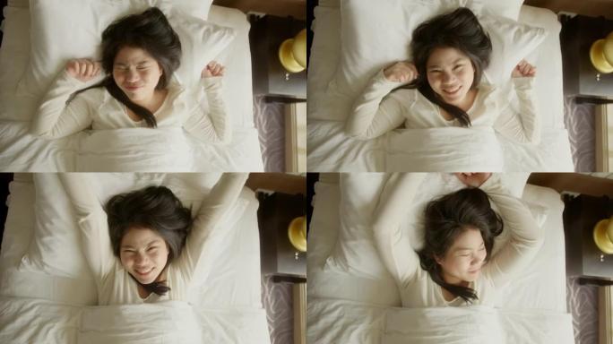 年轻清新的亚洲女女人早上醒来穿着睡衣在床上升起手，在阳光照射下的大窗户旁边，covid锁定结束后的假