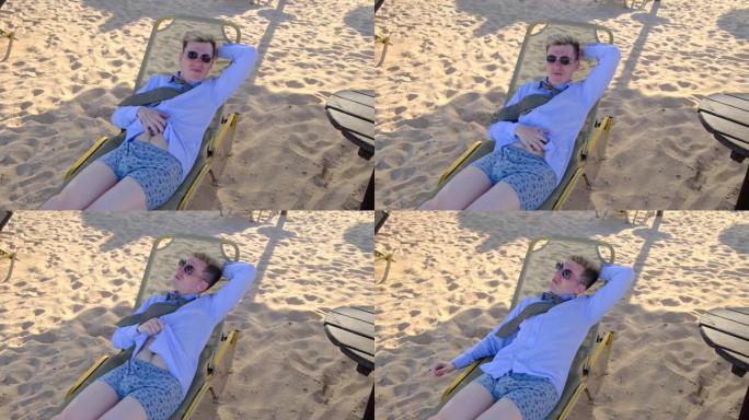 穿着衬衫、领带、内衣和太阳镜的商人抓着肚子，躺在沙滩上的日光浴躺椅上，享受暑假，把手放在脑后，慢动作