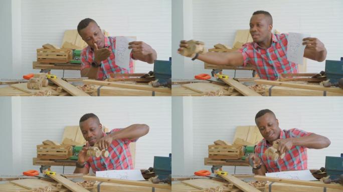 年轻的非洲裔美国男木匠坐在桌子旁，通过平板电脑展示模型车木制和绘图纸，同时与客户在线聊天或在工厂在线
