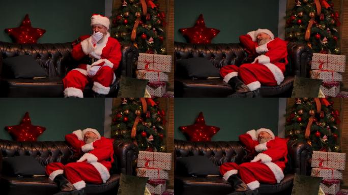 疲倦的圣诞老人打哈欠，坐在沙发上睡着了。老人，留着红色西装的胡须，戴着帽子，在一间发光的圣诞树和装有