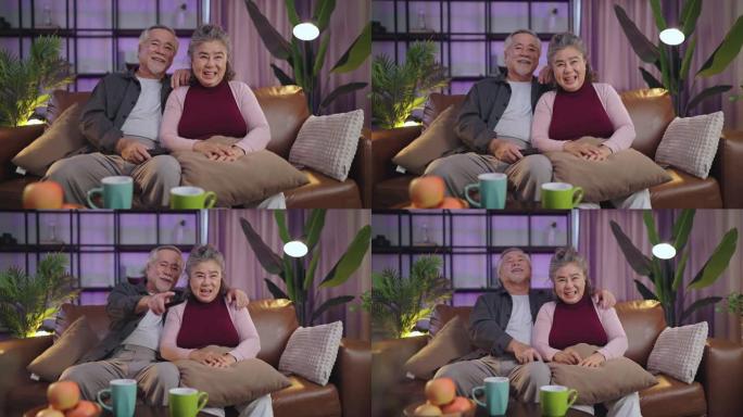 老年退休亚洲夫妇在家看电视，老年成熟亚洲夫妇欢呼体育比赛与笑笑胜利沙发沙发在客厅家庭隔离活动