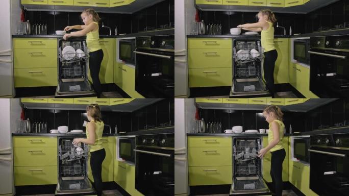 聪明的女孩正在学习使用洗碗机。绿色黑色的现代厨房电器。孩子正在放干净的盘子。