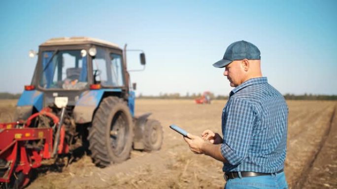 法默正在使用数字平板电脑。在农田上，在拖拉机附近，在农业机械背景上。智能农业。农业技术