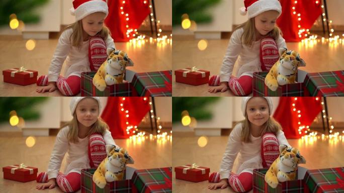 女孩坐在打开的盒子里，在圣诞树旁边玩玩具老虎。新年