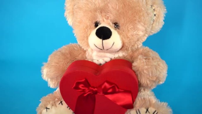 可爱的泰迪熊的特写镜头，红色礼品盒呈心形。蓝色背景的毛绒玩具。假期、礼物和好心情的概念