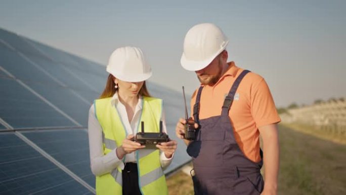 技术人员和投资者使用红外无人机技术检查太阳能电池农场中的太阳能电池板。戴着安全帽的工程师妇女拿着平板