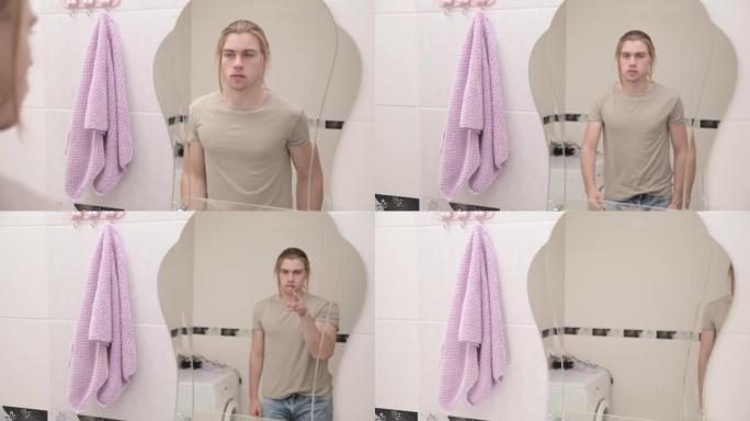 一个年轻的长发男人走到浴室的镜子前，早上看着自己。离开画面显示他在跟踪摄像机
