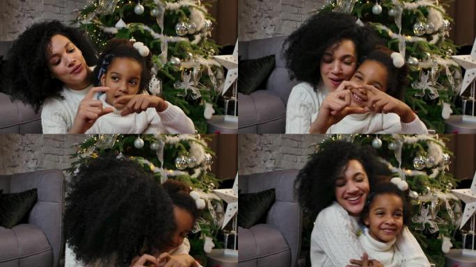 快乐微笑的非洲裔美国妈妈和小女儿看着相机，用手指做心脏手势的肖像。家庭在装饰圣诞树的背景。特写。慢动