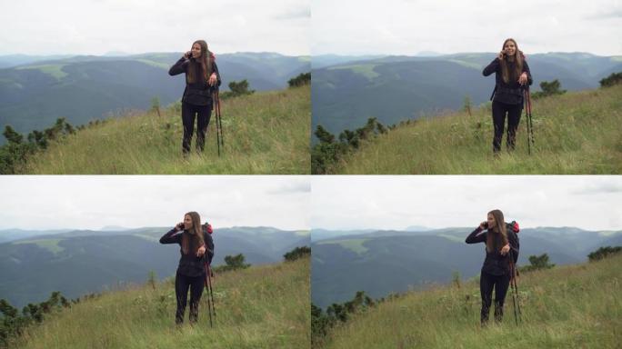 一个背着背包的年轻女子站在秋天或春天的山顶上，拿着拐杖，用智能手机与人交流。徒步旅行概念。