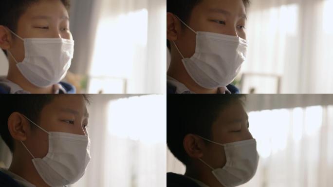 亚洲男生早上在上学前在家窗户附近戴口罩，这是新的常态和社交疏远的生活方式理念。