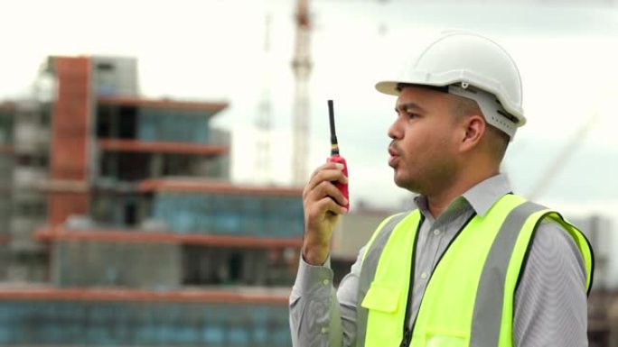 亚洲工程师帅哥或建筑师在城市建筑工地使用带有白色安全帽的无线电通信。站在首都的屋顶建筑上。
