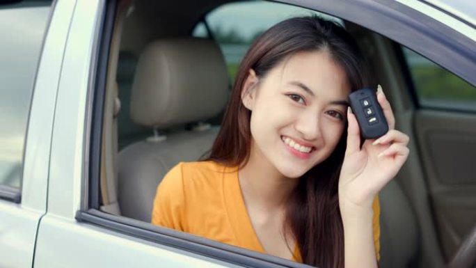 年轻漂亮的亚洲女性得到新车。她手里拿着车钥匙看着窗外，非常高兴和兴奋。灿烂的日子里，微笑的女性驾驶车