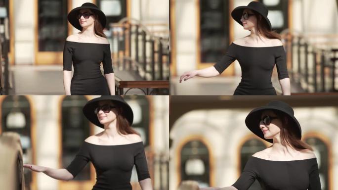 穿着黑色连衣裙、帽子和太阳镜的时尚迷人女士走在商场的街道上。经典时尚概念。慢动作