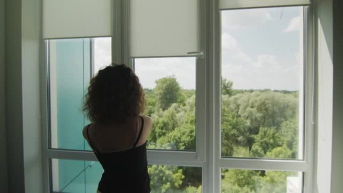 卷发的年轻女孩的后视图，拉起窗户上的织物百叶窗。阳光明媚的夏日