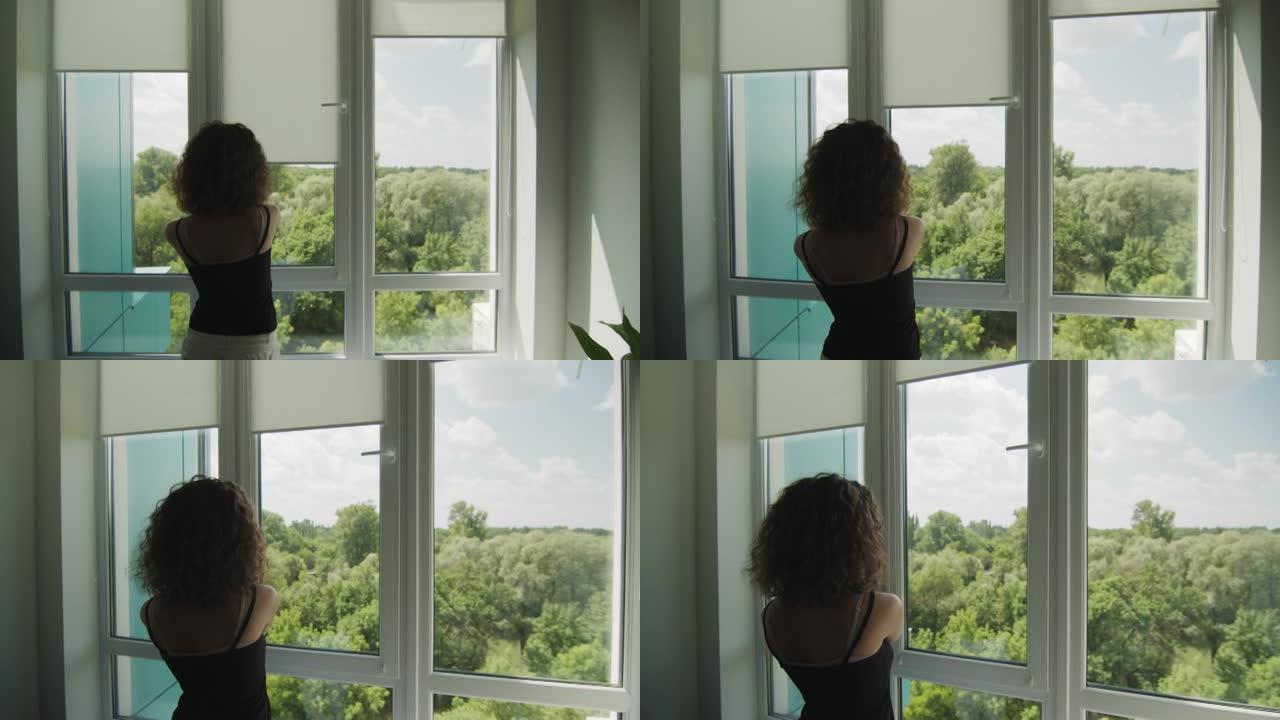 卷发的年轻女孩的后视图，拉起窗户上的织物百叶窗。阳光明媚的夏日