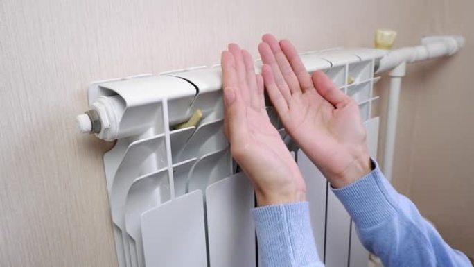 一名女性在房间里家里用电池加热。手指，手掌从散热器中保持温暖，关闭。室温低。冬季采暖期，季节。