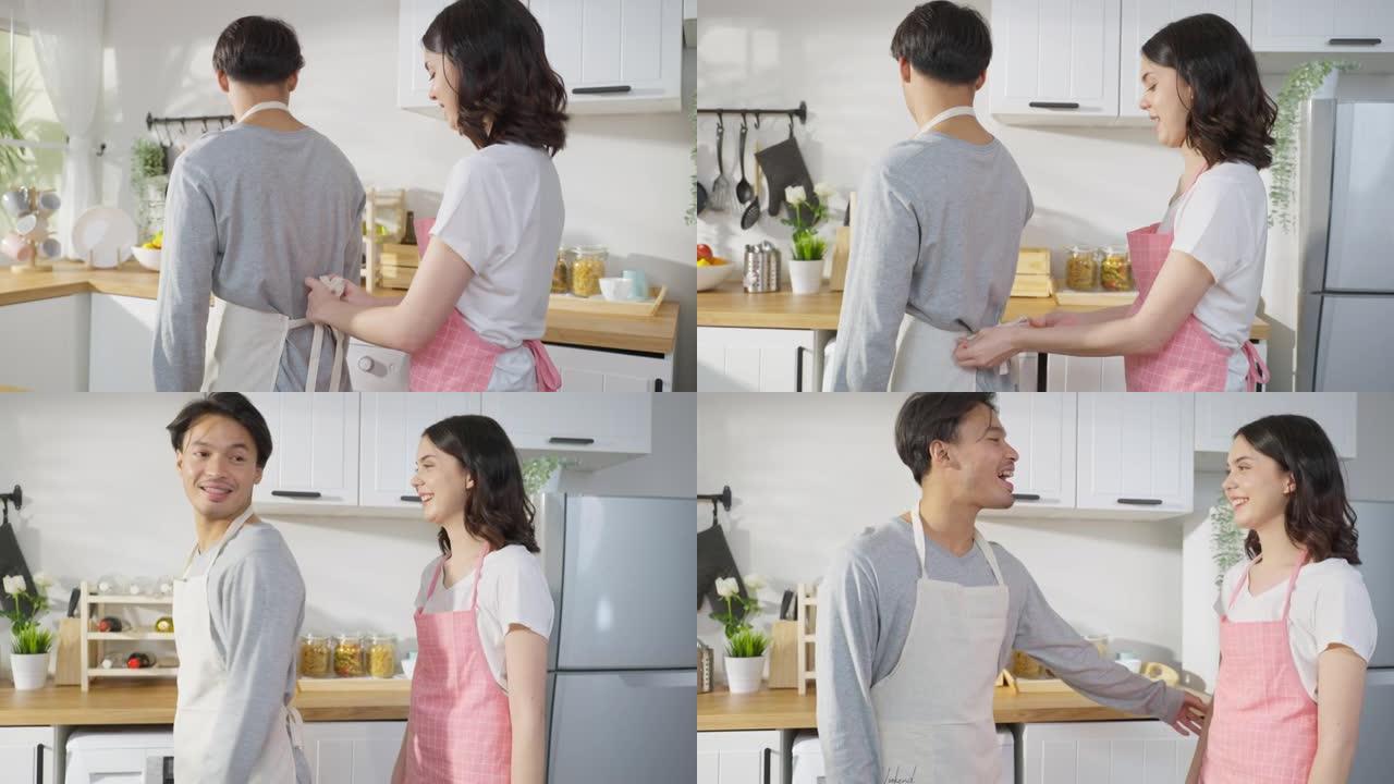 亚洲年轻新婚妻子在厨房给丈夫绑围裙鞋带。迷人的浪漫情侣穿着围裙在家里共度时光，感到快乐，喜欢在家里烹