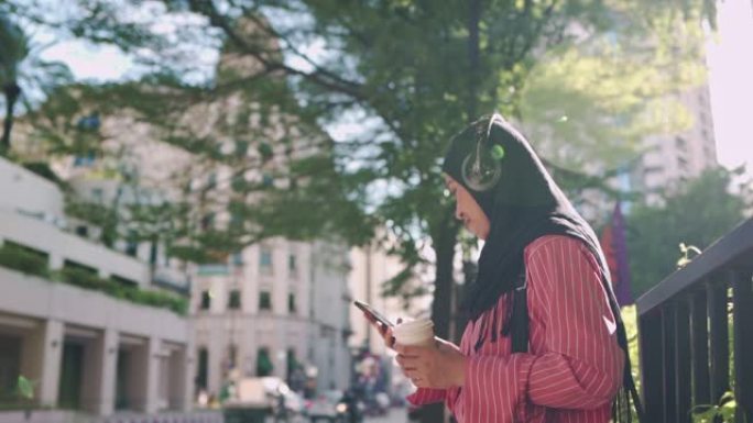 亚洲穆斯林女性戴着耳机听播客，温暖阳光明媚的日子，拿着咖啡杯站在街边下午咖啡休息时间，使用智能手机，