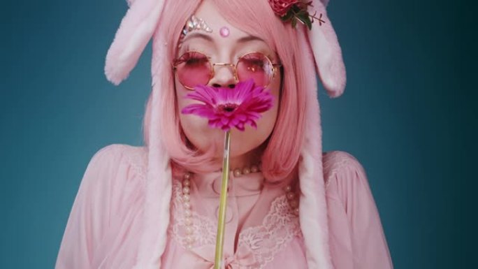 快乐的粉红色可爱的糖果动漫女孩与兔子耳朵和粉红色太阳镜的特写镜头嗅着一朵花和蓝色背景上的情感鬼脸。美