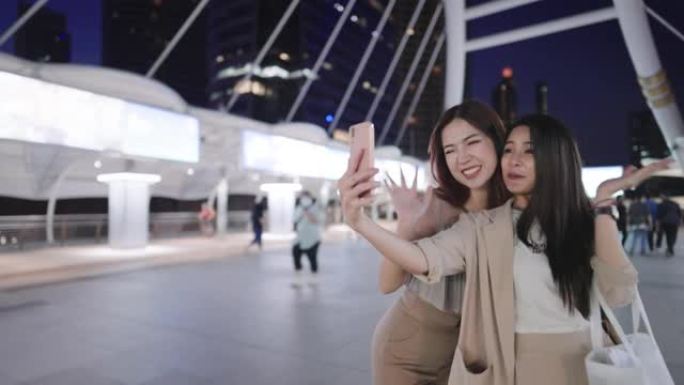 两位兴高采烈的亚洲游客对着智能手机和挂起远方朋友的视频电话，快乐时尚的上班族们一起在著名城市大桥的夜