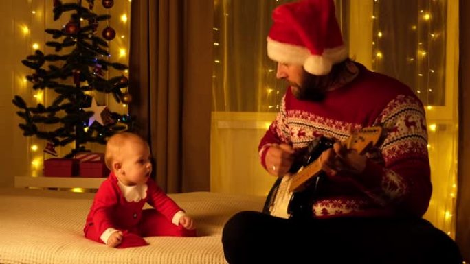 父亲戴着圣诞老人的帽子和毛衣给他的小孩弹吉他。宝贝看着他，听着，微笑着。圣诞灯背景。圣诞节快乐家庭概