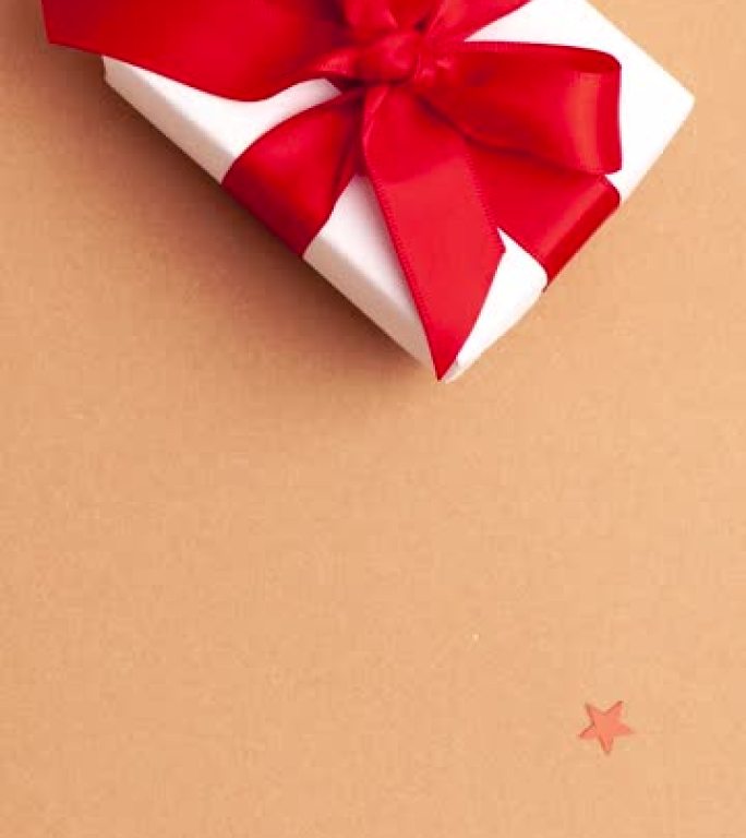 穿着白色毛衣的人的手拿走了一份白纸礼物，上面有一个红色缎带蝴蝶结，棕色背景是红色星星。停止运动垂直动