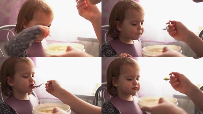 食物，营养，童年概念-中等射击妈妈用橡胶勺子喂养婴儿。可爱的饥饿的孩子穿着围兜坐在孩子们的椅子上，不