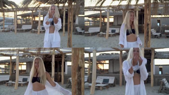 在一个热带度假胜地，一位美丽的金发女子在沙滩上的草伞之间向前走去，看着相机，用手招手。