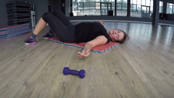 超重的女人躺在健身俱乐部的地毯上很累扔掉哑铃不能呼吸，从内啡肽高兴