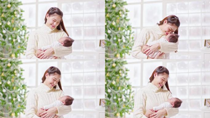 美丽的亚洲女人抱着新生婴儿，亲吻坐在白色舒适的家中，背景是圣诞树。快乐的婴儿睡在母亲的怀抱中安全舒适