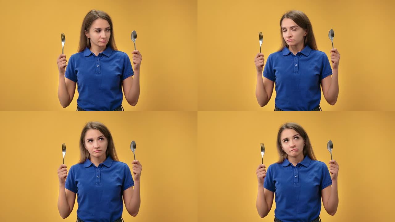 肖像悲伤的女人在饮食中选择叉子和勺子在橙色上挨饿