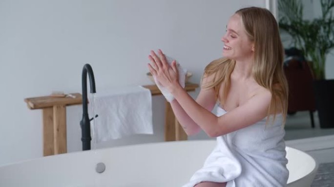 一个金发碧眼的漂亮女人，裹着白色毛巾布，坐在浴缸边上，试着水温，用肥皂泡澡。清新、纯净、呵护身体的理