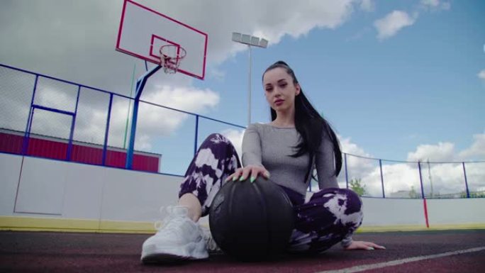 迷人的黑发女篮球运动员坐在黑球旁边，看着篮球室外球场背景上的相机，白天