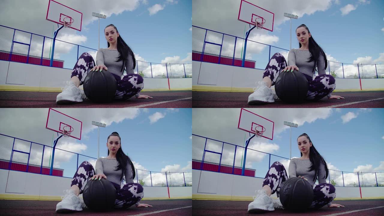 迷人的黑发女篮球运动员坐在黑球旁边，看着篮球室外球场背景上的相机，白天