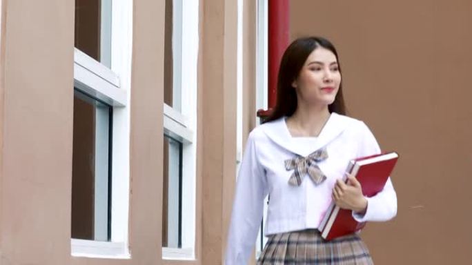 穿着日式大学制服的可爱的年轻女学生拿着书，带着快乐的脸走在shcool大楼前。