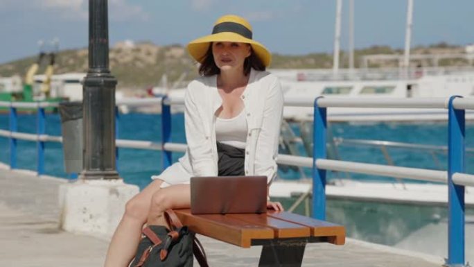 一个戴着帽子、拿着笔记本电脑的成熟女人在城市的海边散步。