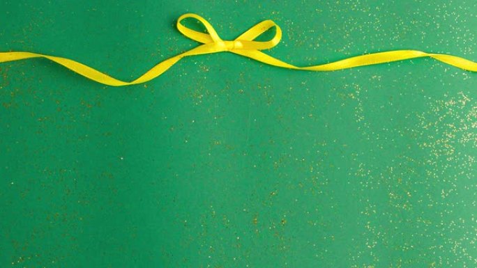 黄色缎带蝴蝶结绑在玉绿色背景上，金色闪光。定格动画圣诞假期和呈现概念平铺与复制空间