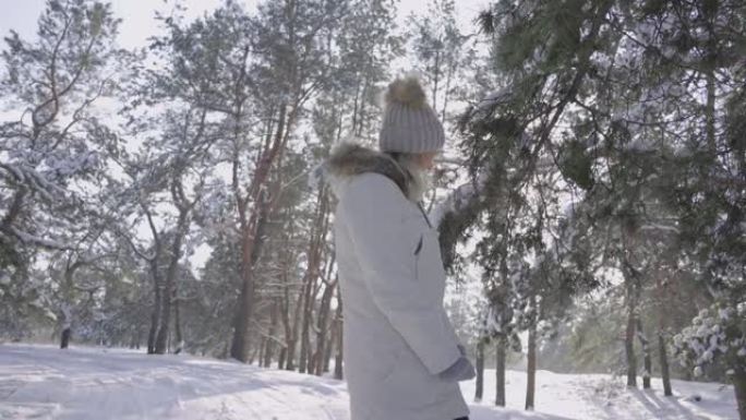 一个穿着冬装的漂亮女孩开心地笑着，享受着寒冷的晴天，呼吸着新鲜的空气。走在白雪皑皑的森林里。松枝上有