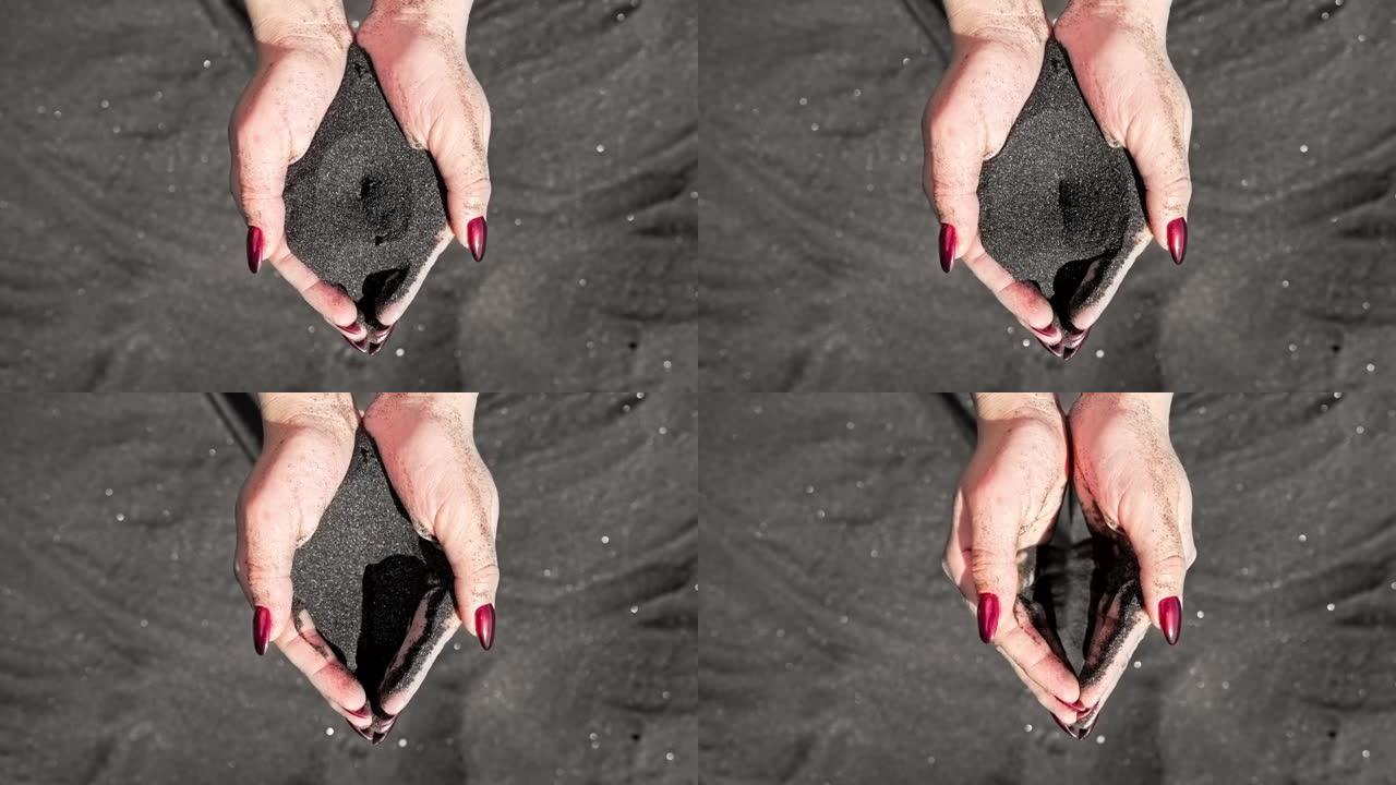 在岛屿热带海滩上，女性的手在慢动作中倒入火山黑沙，穿过手指。美丽的场景，年轻女子与红指甲玩沙砾在日落